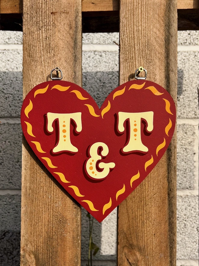 Wooden love heart cut out with a red matte background, glossy golden yellow curve details painted along the edges and ivory lettering with dark red shade. It is hanging on a wooden pallet.