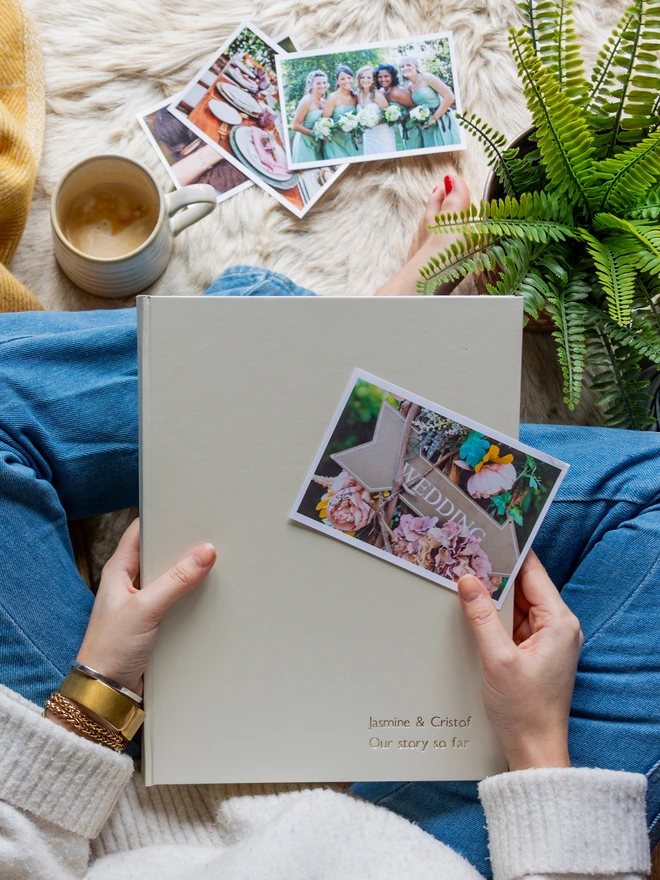 A woman in a pair of jeans is sitting cross legged on a cosy rug with a cup of coffee and a white A4 sized photo album.