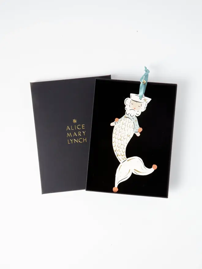 Merman Tree Dancer. the merman tree dancer hanging ornament sits inside its black presentation box, with the lid open to reveal "alice mary lynch" embossed in gold lettering. the bearded merman figure wears a sailor hat and striped teal top, with a cream scale-patterned tail edged in gold and dotted with silver, curling into a jester-style pointed tip with orange-tipped ends. the branded box gives the piece a polished, gift-ready presentation.