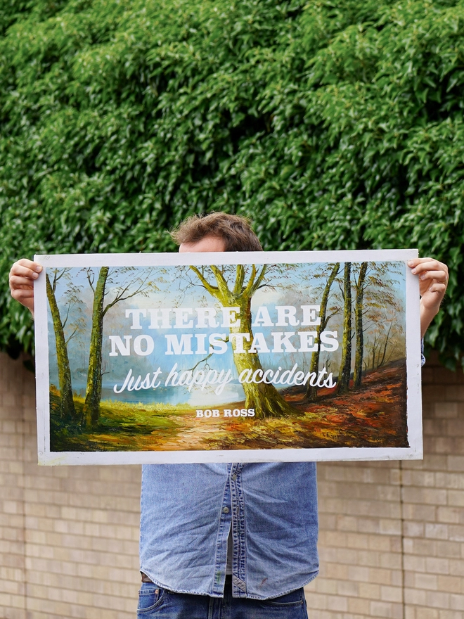 photograph of a man holding a large original oil painting depicting a woodland scene with the words 'there are no mistakes, just happy accidents' Bob Ross screen printed over the top in white ink