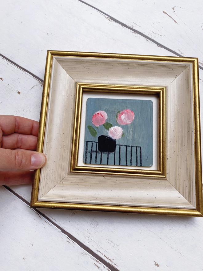hand holding picture of three painterly pink flowers on dusty blue background in a cream and gold 1960's style frame