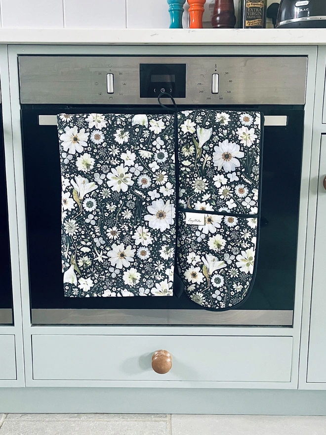 Floral double oven gloves and tea towel with pressed white flower design on a black background. Complete with hanging loop.