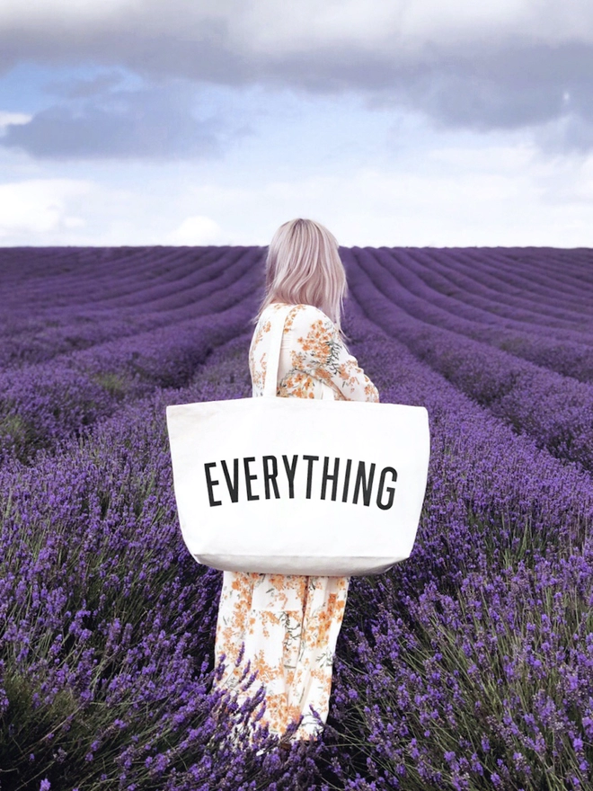 Model standing in a lavender field carrying the Everything oversized tote bag in natural canvas