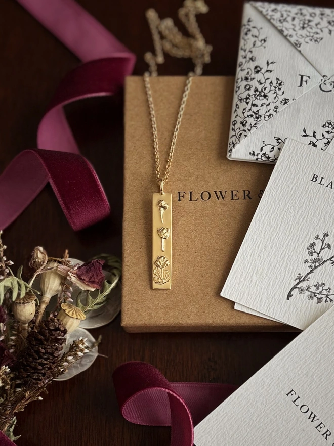 Close up of a Gold bar pendant belcher necklace representing luck draped over flower & fable packaging