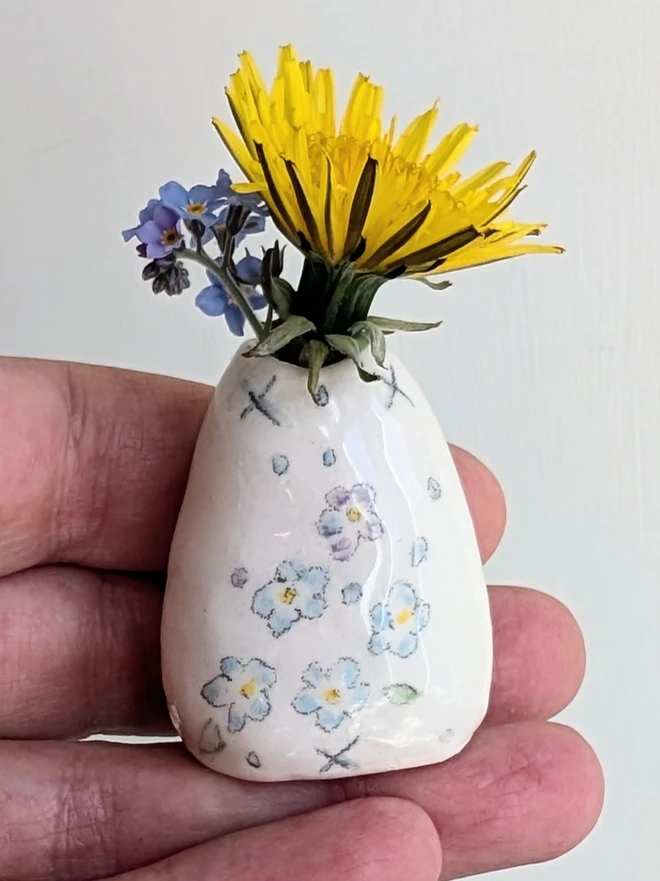 forget me not miniature vase held in a hand with dandelion and forget me not flowers in and hand painted illustration