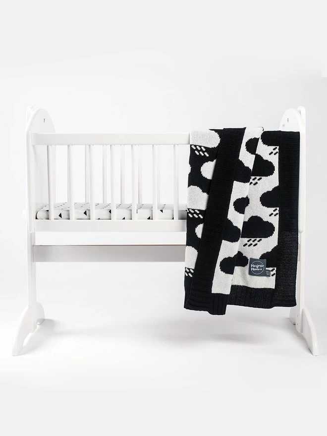 A black and white storm cloud design blanket is draped over the side of a baby's cot.
