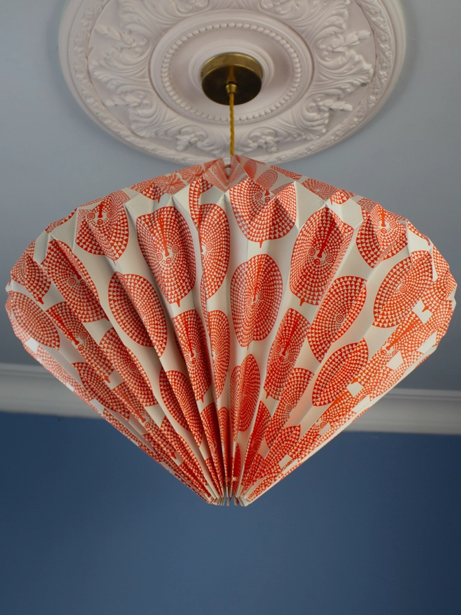 A pendant light with an orange and white geometric patterned shade hangs from an ornate ceiling rose against a blue wall.