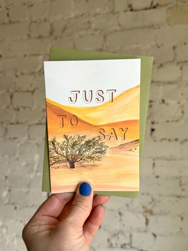 a greetings card featuring an illustration of a desert landscape with the text “just to say” with a green envelope