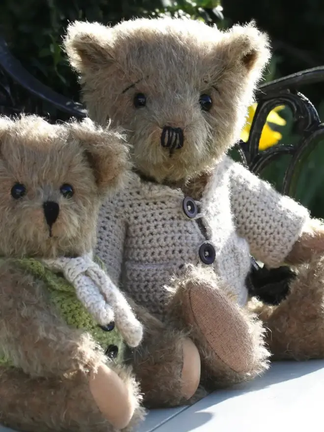 Brown teddy bear wearing a cream knitted cardigan sitting among a group of other teddy bears.