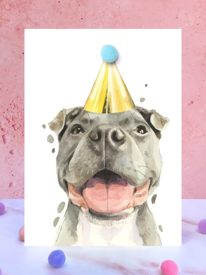 A greeting card featuring a hand painted design of a staffordshire bull terrier, stood upright on a marble surface surrounded by pompoms. 