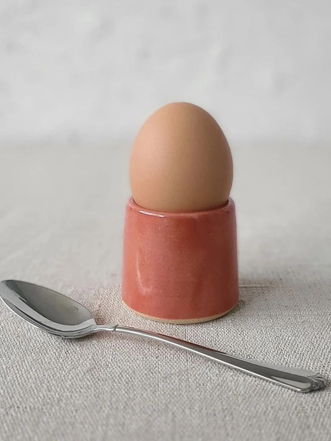 Classic Handmade Egg Cup, a ceramic egg cup sitting on a carpet surface against a grey backdrop. It is accompanied by a spoon. 