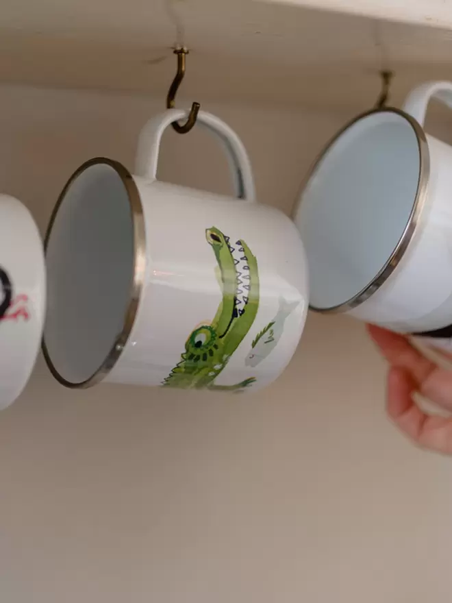 White enamel mug with green corcodile decoration, hanging from a hook under a shelf