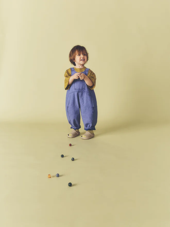Overhead crop of a toddler holding a handful of marbles, wearing Studio Koter washed blue denim Kai dungarees over an ochre t-shirt.
