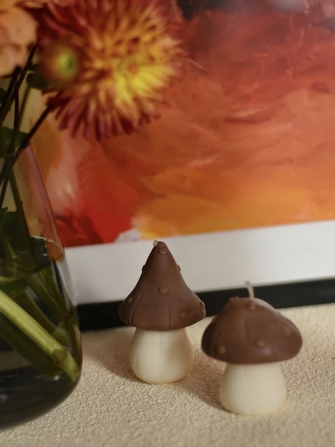 Pair Of Little Mushroom Candles (Set Of 2). Two mushroom candles with chocolate brown dotted caps and cream stems, one with a pointed cap and one rounded, set beside a vase of dahlias and a colourful painting. The playful handmade pair makes a charming accent for a shelf or side table.