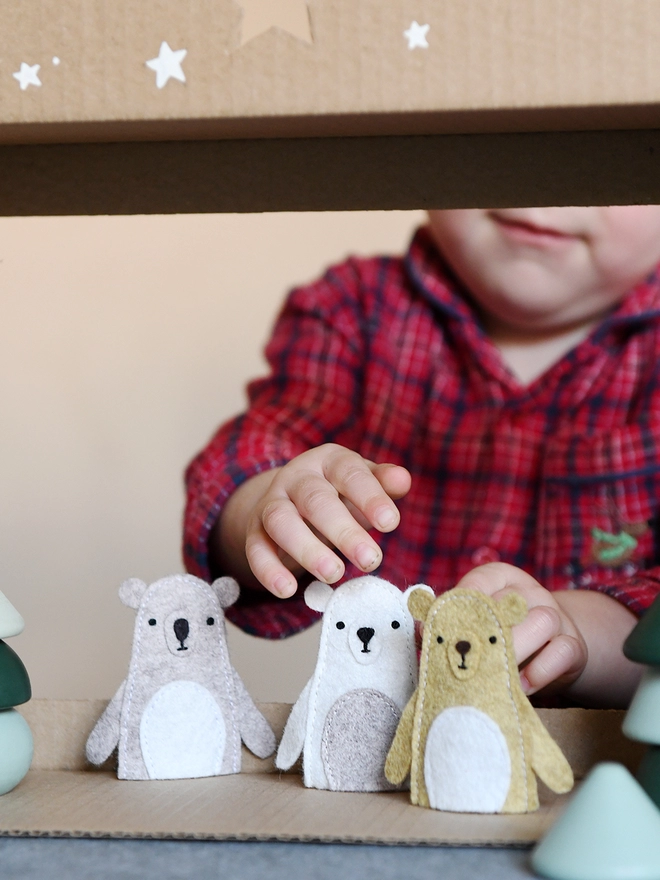 A young child wearing red pyjamas holds three handmade felt bear finger puppets on a cardboard puppet theatre.