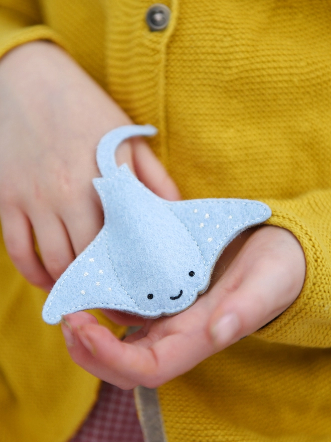 A young child wearing a yellow cardigan is holding a soft blue felt manta ray finger puppet on their fingers.