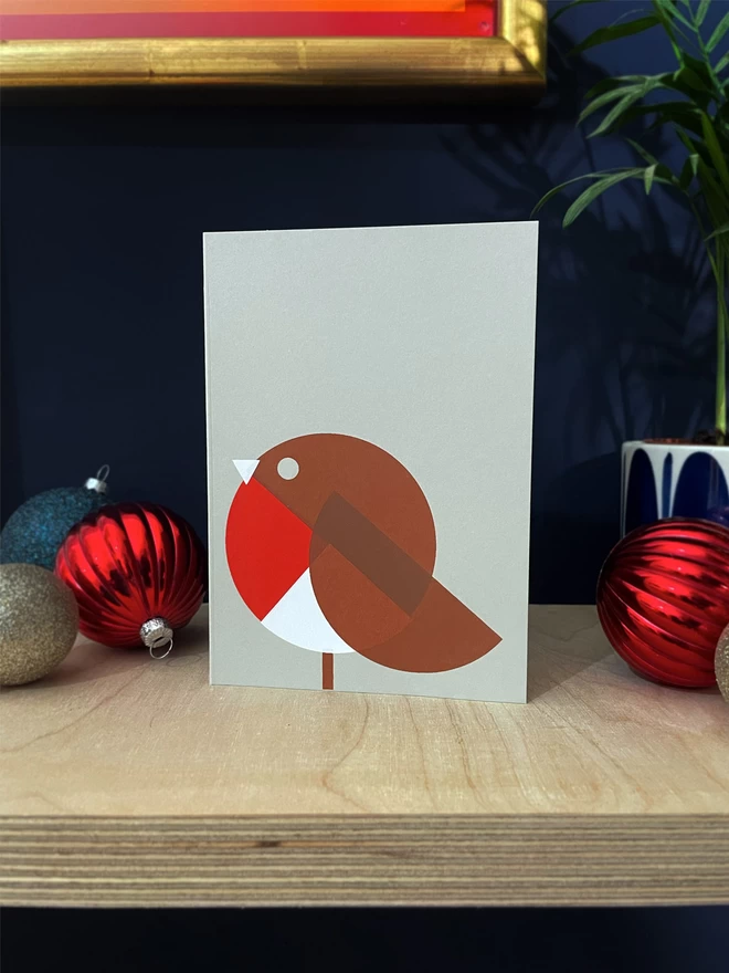 Geometric shapes in red white and brown make up this handprinted robin christmas card, stood on a plywood shelf with baubles and foliage 