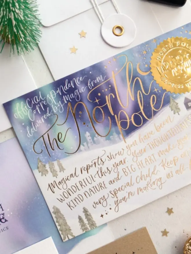 An angled close-up of the northern lights North Pole notecard, showing the gold foil "The North Pole" calligraphy, the "Official correspondence delivered by magic from" heading and the embossed gold seal against a white string-and-washer envelope.