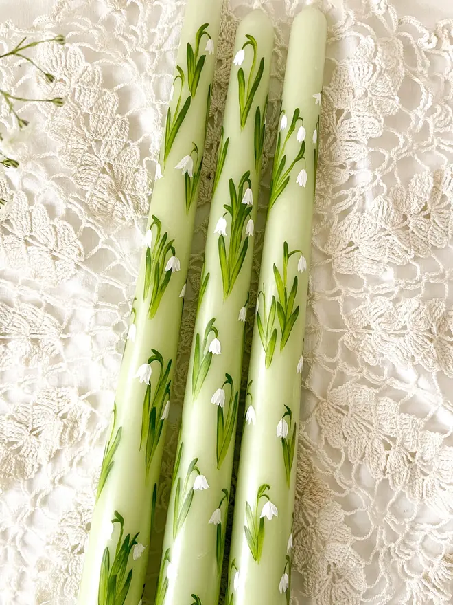 Snow Drops Handpainted Candle, a group of snowdrop candles lying on a white table. They are accompanied by the snowdrop flower. 