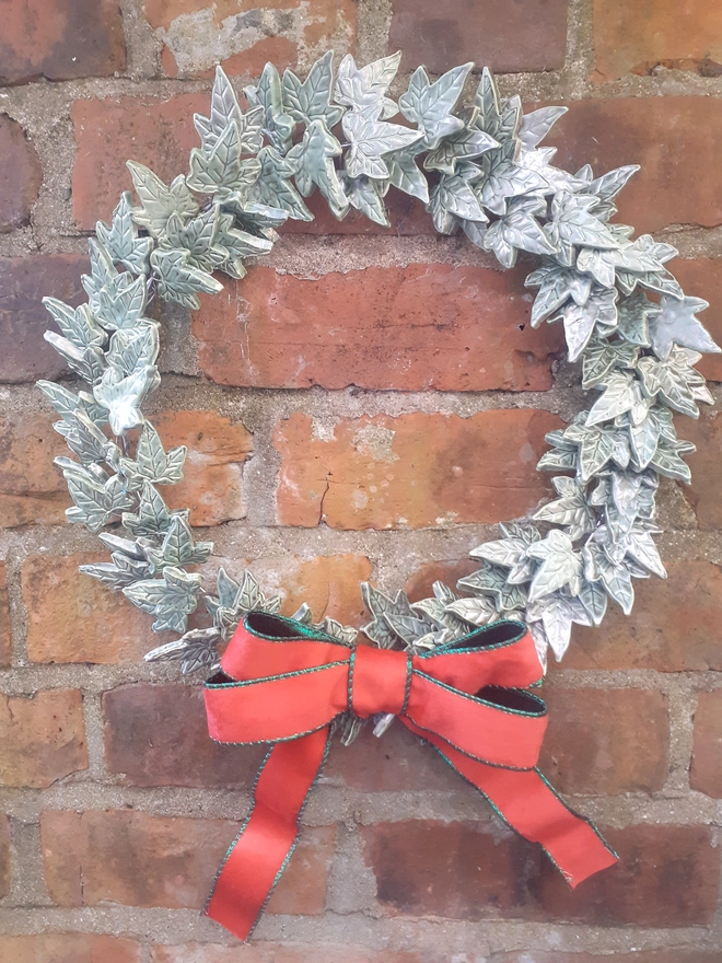 Handmade ceramic ivy leaf wreath against a rustic brick wall, glazed in verdigris green with metallic copper highlights and finished with a large red velvet bow.