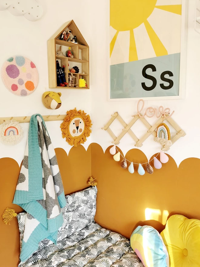A modern childrens room with a cosy cushioned reading corner and half scalloped ochre walls. The walls are adorned with fun childrens decor, and on a wall peg hangs a grey and white knitted cloud baby blanket with a mint green trim.