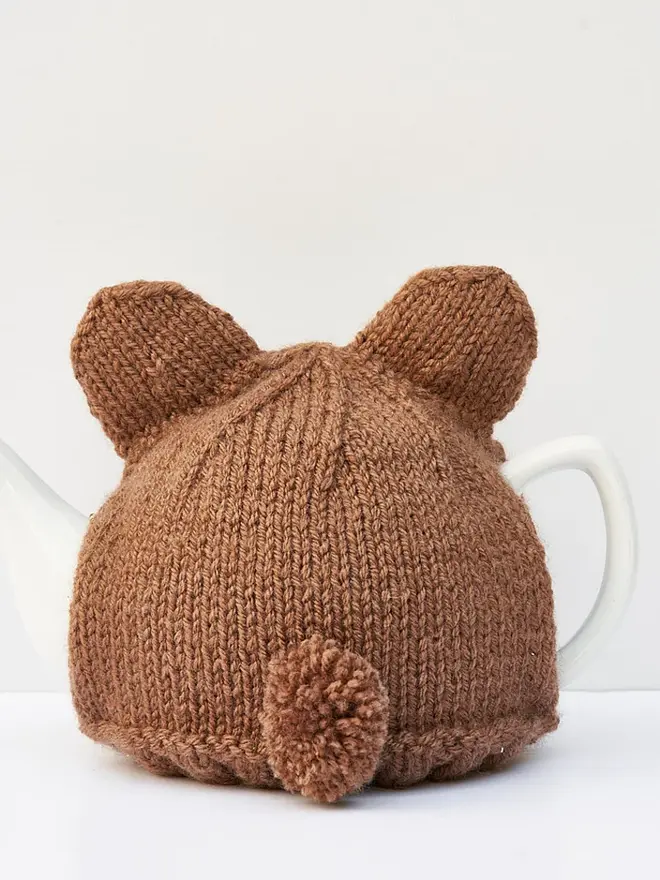 Brown Bear Tea Cosy Teddy Bears Picnic Treat, a brown bear tea pot cover sitting on a white tea pot, on a white surface against a grey backdrop. 