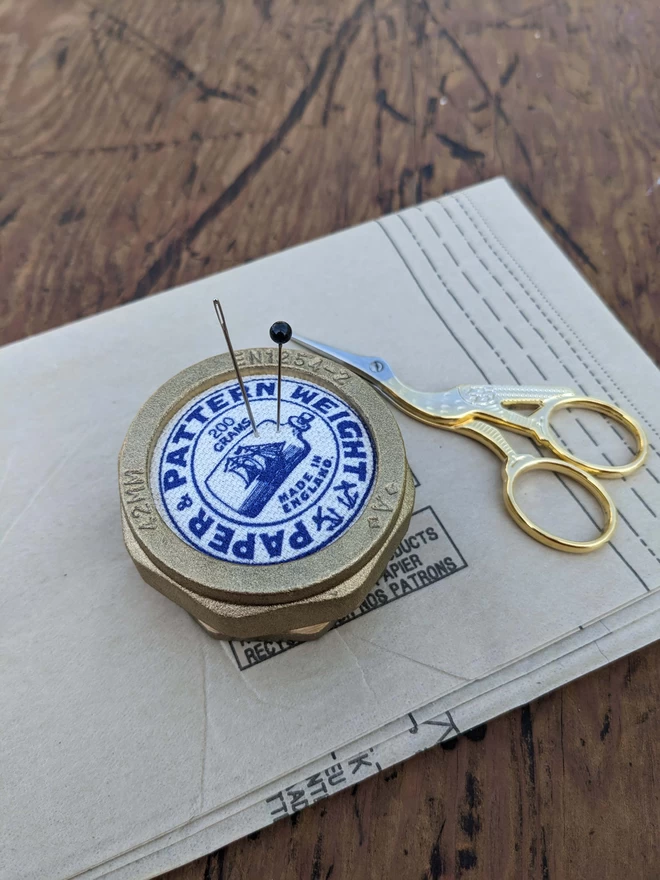 Blue pattern and paper weight on its side holding a needle and pin on vintage sewing pattern paper next to embroidery scissors