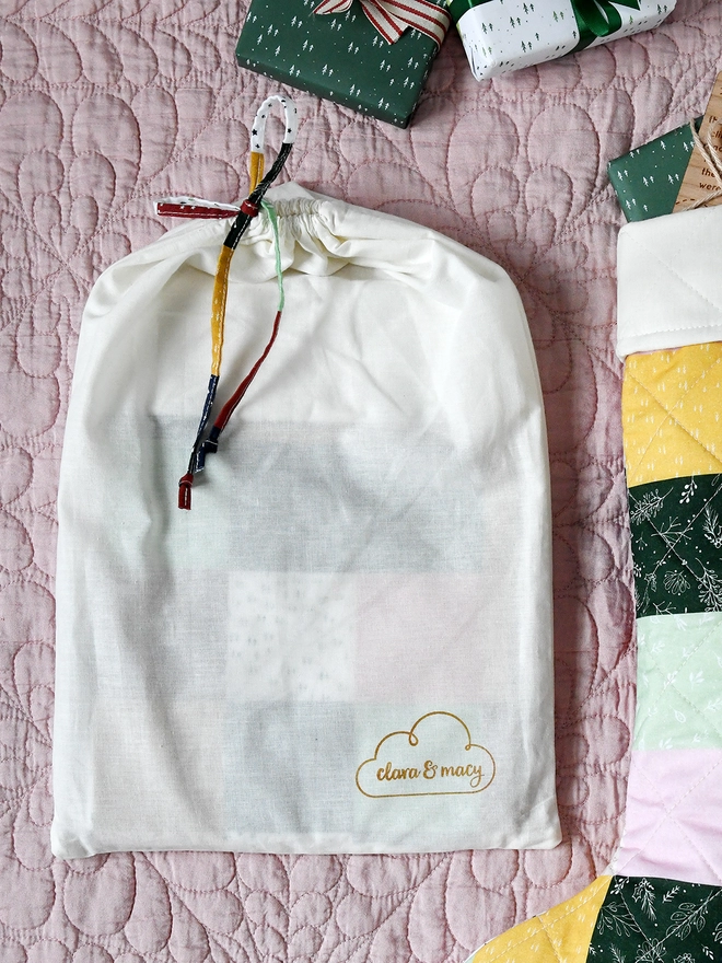 An ivory cotton dust bag lays beside a patchwork stocking on a pink quilt.