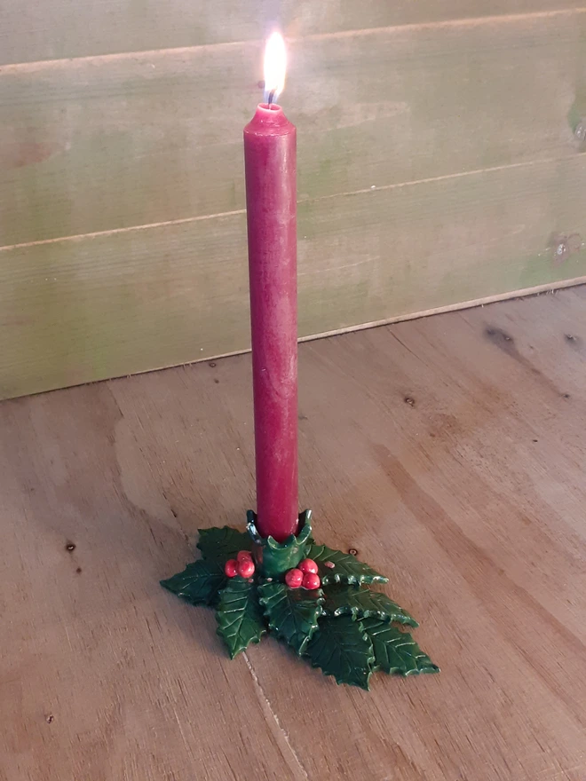 Red dinner candle glowing in a handmade ceramic holly leaf holder with glossy green leaves and red berries, styled on a rustic wooden background.