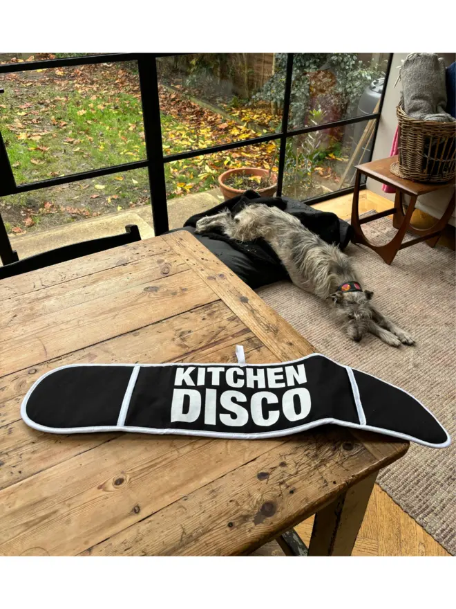 Black oven glove that says KITCHEN DISCO on it laying on a wooden table in front of a large window with a dog asleep on the floor behind it in the background 