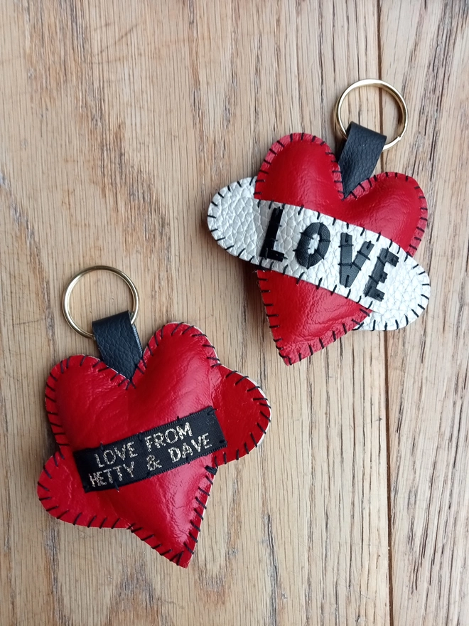 A red heart keyring which says LOVE in black lettering across a white scroll, along with the red leatherette reverse of a heart keyring, showing the black label with LOVE FROM HETTY & DAVE in gold lettering