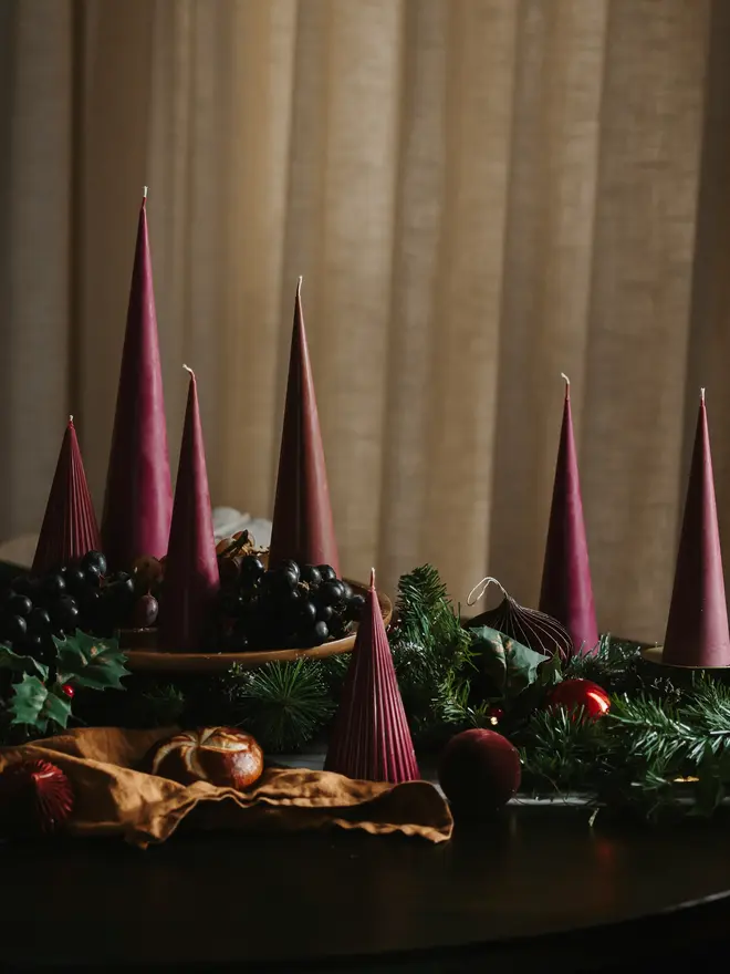 Burgundy Christmas Tree Cone Candles (Set Of 3). Burgundy cone candles in a range of heights, some smooth and some finely ribbed, set among black grapes, pine garland, holly and velvet baubles on a dark table. The rich berry tone makes them a striking, sculptural centrepiece for Christmas dinner.