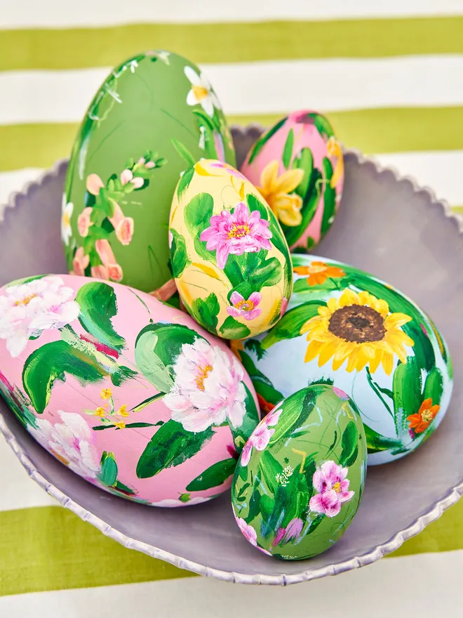 Mixed colours of hand painted wooden eggs positioned in a purple scalloped bowl, atop a green and white striped tablecloth