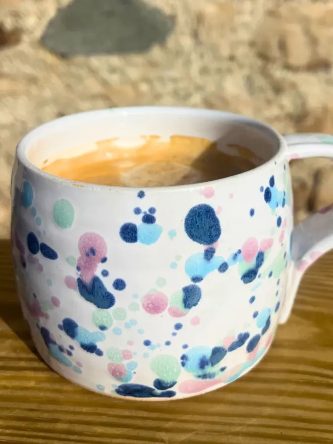 Sweetpea Large Round Mug, a colourful mug sitting on a wooden surface against a brick backdrop.