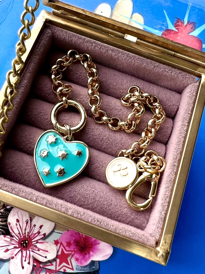 Gold belcher chain bracelet with a turquoise enamel heart charm set with tiny gold stars inside a pink velvet jewellery box
