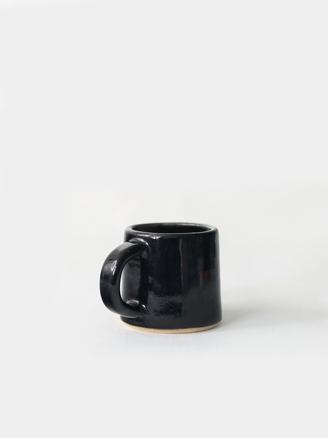 A handmade ceramic espresso mug in jet black glaze, with a chunky loop handle, photographed against a plain light grey background