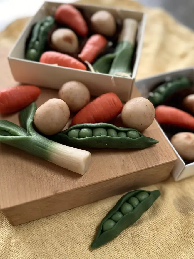 Veggilicious Marzipan Box, a marzipan vegetable box on a wooden chopping board on a yellow table cloth. In the box are marzipan peas, spring onions, potatoes and carrots. 