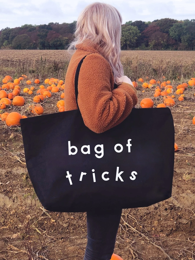 model holding out an oversized black canvas tote bag with the words bag of tricks in a pumpkin patch at halloween