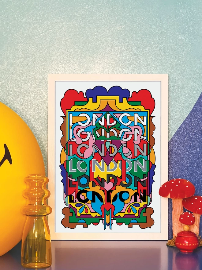 A vibrant, portrait print containing all the colours of the London Underground lines, with the word London repeated six times from top to bottom. The picture is in a white frame leaning against a painted blue wall on a blue cabinet. Next to the frame is a large illuminated yellow Smiley lamp, a yellow vase, a model of three red and white toadstools and a small round red wooden pot.