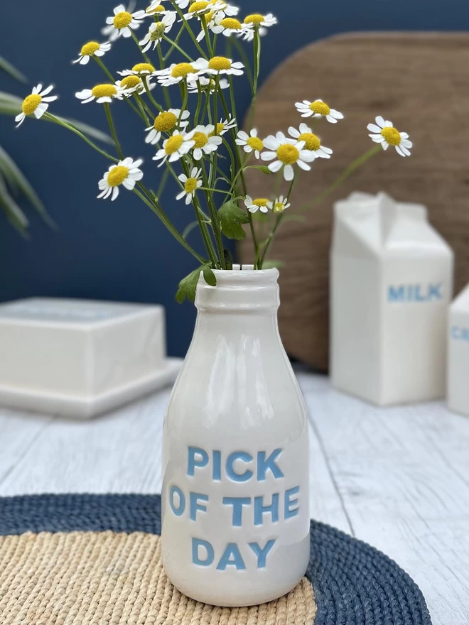 A handmade ceramic bottle/vase with ‘pick of the day’ lettering painted in blue, holding flowers.