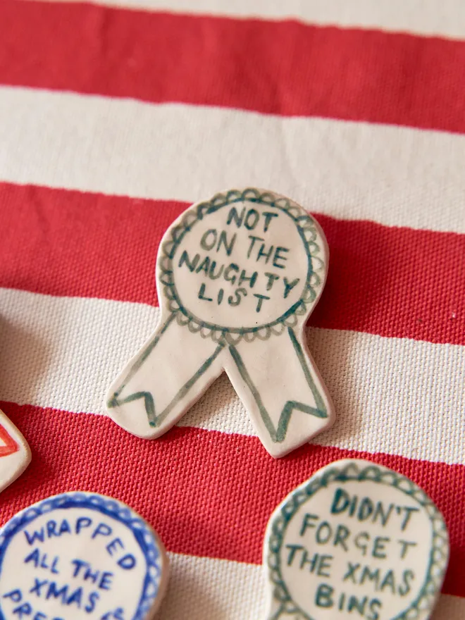 A selection of Red, Green & Blue Handmade Ceramic Christmas Award Badges, handpainted with different phrases, atop a striped red and white tablecloth