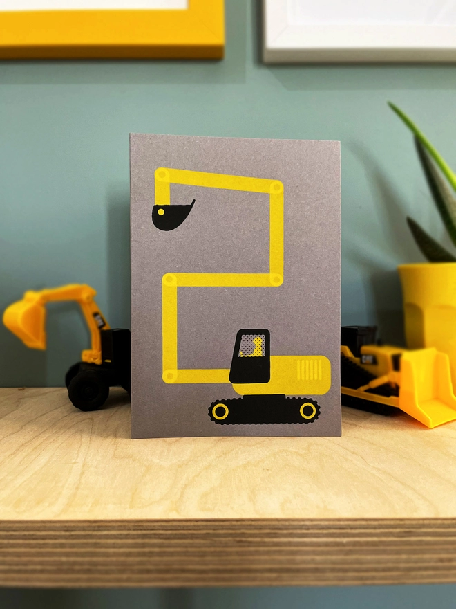 A digger making a number 2 with its digger arm, screenprinted in yellow and black ink on a grey card. on a plywood shelf with little digger toys besides and other framed pictures around.