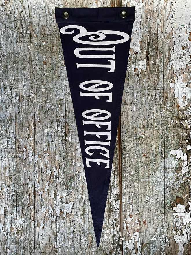 A navy canvas pennant flag hung vertically on a wooden wall with the words 'Out od Office'  written in white canvas.
