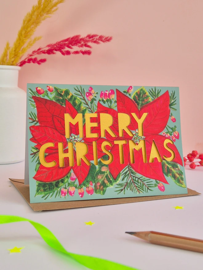 Red Poinsettia Illustration Christmas Card with green background, paper cut text that says Merry Christmas and has a pearlescent gold backing paper 