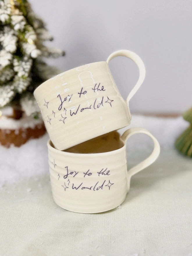 a stack of two cream hand thrown porcelain mug with the words 'Joy to the World' printed onto the side with pretty line drawn stars. Sitting on a tabletop with snow, a mini lit Christmas tree 