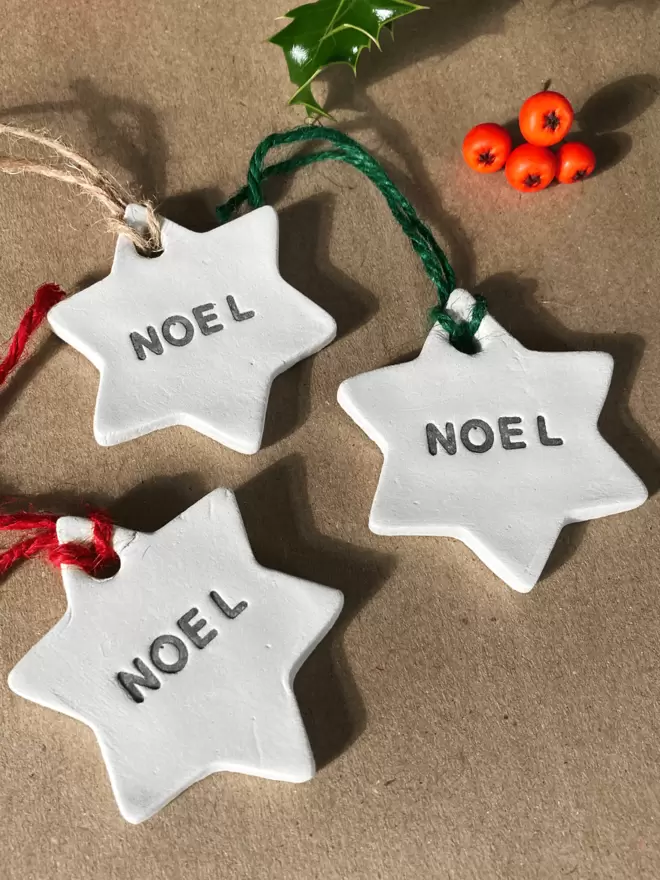 3 star shaped clay tags with 'NOEL' stamped in the centre of the star. Each finished with either red, brown or green twine. Brown paper parcel background with a holly and berries in the top half of the image.