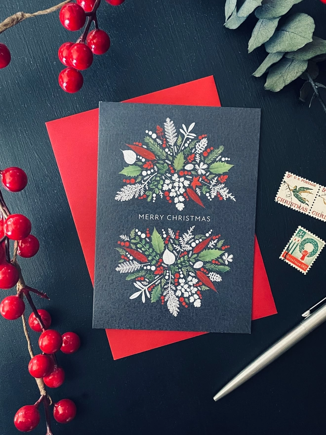 Christmas card with intricate pressed Holly and Ivy leaf design on dark charcoal desk with red berries, eucalyptus, Christmas stamps and pen