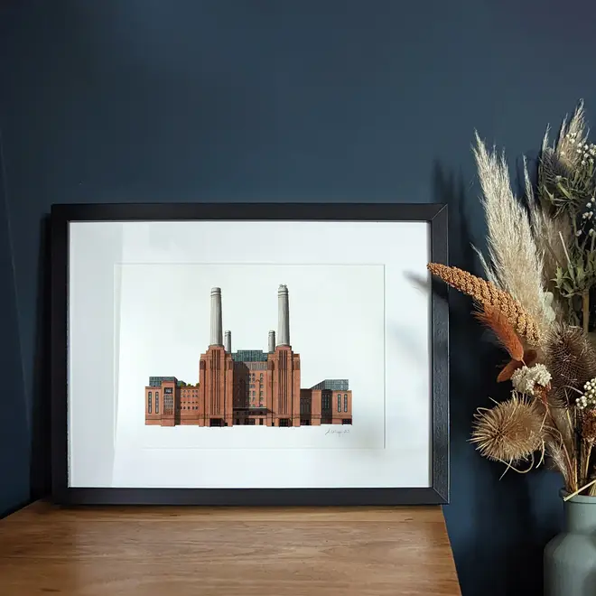 Battersea Power Station Giclée Print (Unframed), a print of Battersea Power Station sitting in a black frame against a navy blue backdrop on a wooden surface. It is joined by a plant. 