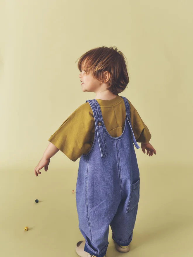 Back view of a toddler looking over their shoulder against a pale yellow backdrop, showing the straps and rear patch pocket of Studio Koter washed blue denim Kai dungarees, worn over an ochre t-shirt.