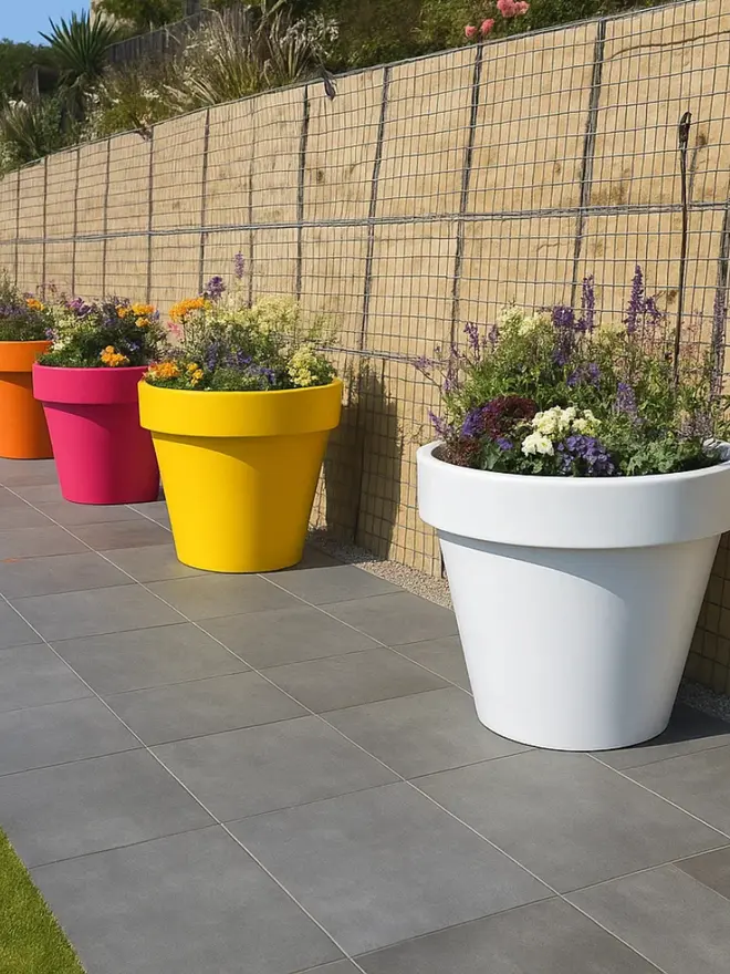 A row of large, modern Biggie plant pots in bright colours: white, yellow, pink, orange, green and blue each filled with flowering plants. adding a pop of bright colours to this garden.